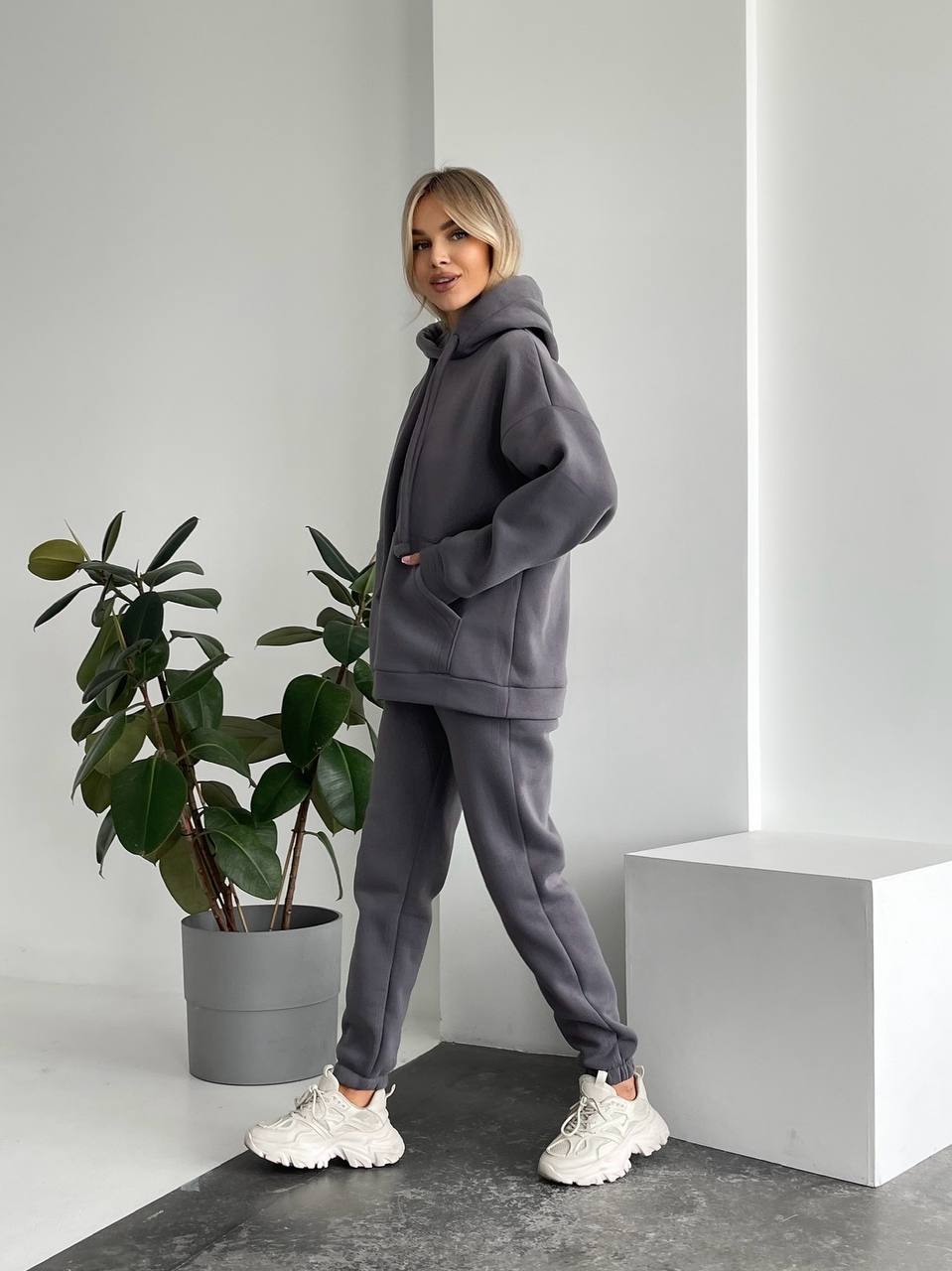 Person wearing a gray tracksuit standing next to a potted plant in a minimalistic setting