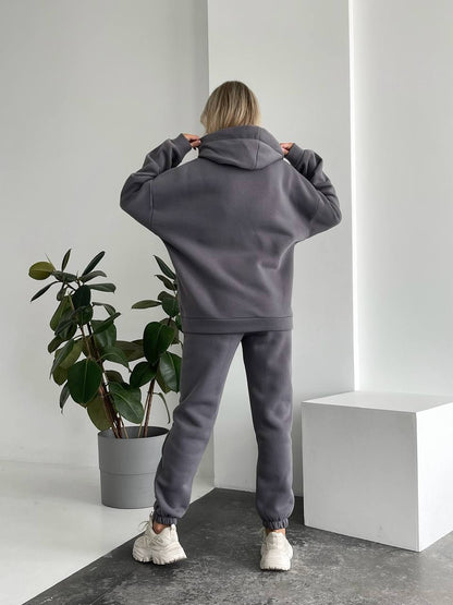 Person wearing a gray tracksuit standing in a minimalistic room with a plant and white cube.