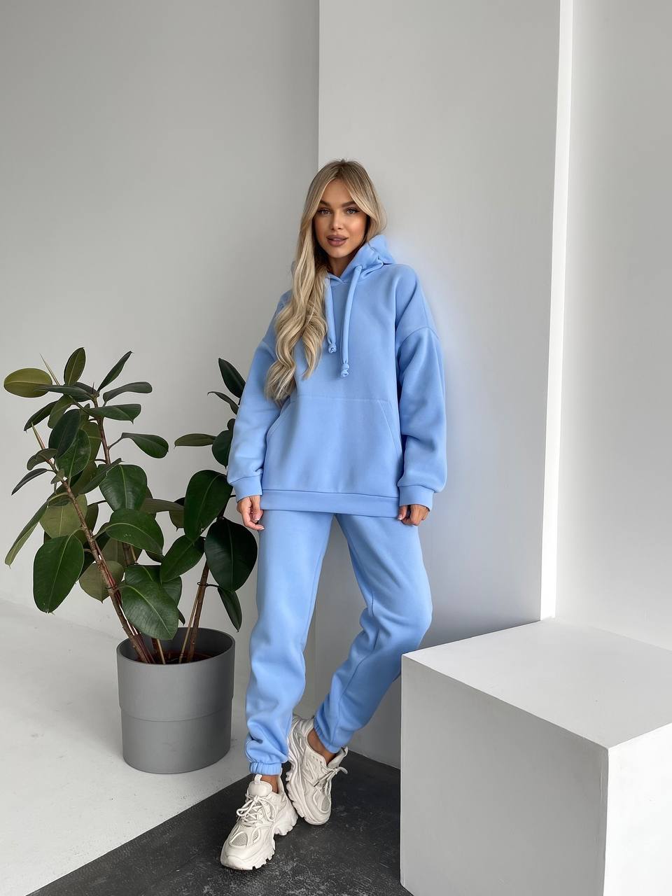Person wearing a light blue tracksuit standing next to a potted plant indoors.
