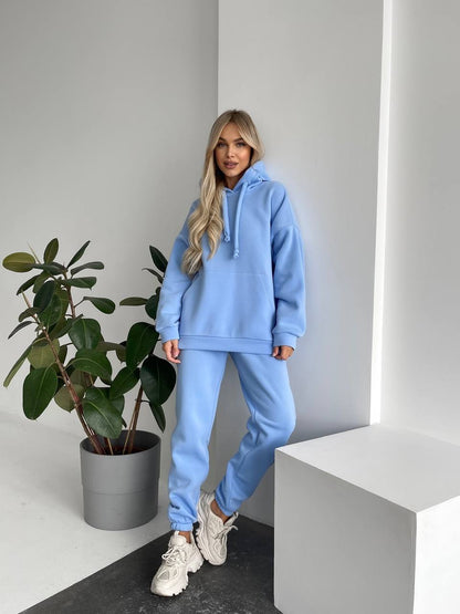 Person wearing a light blue tracksuit standing next to a potted plant indoors.