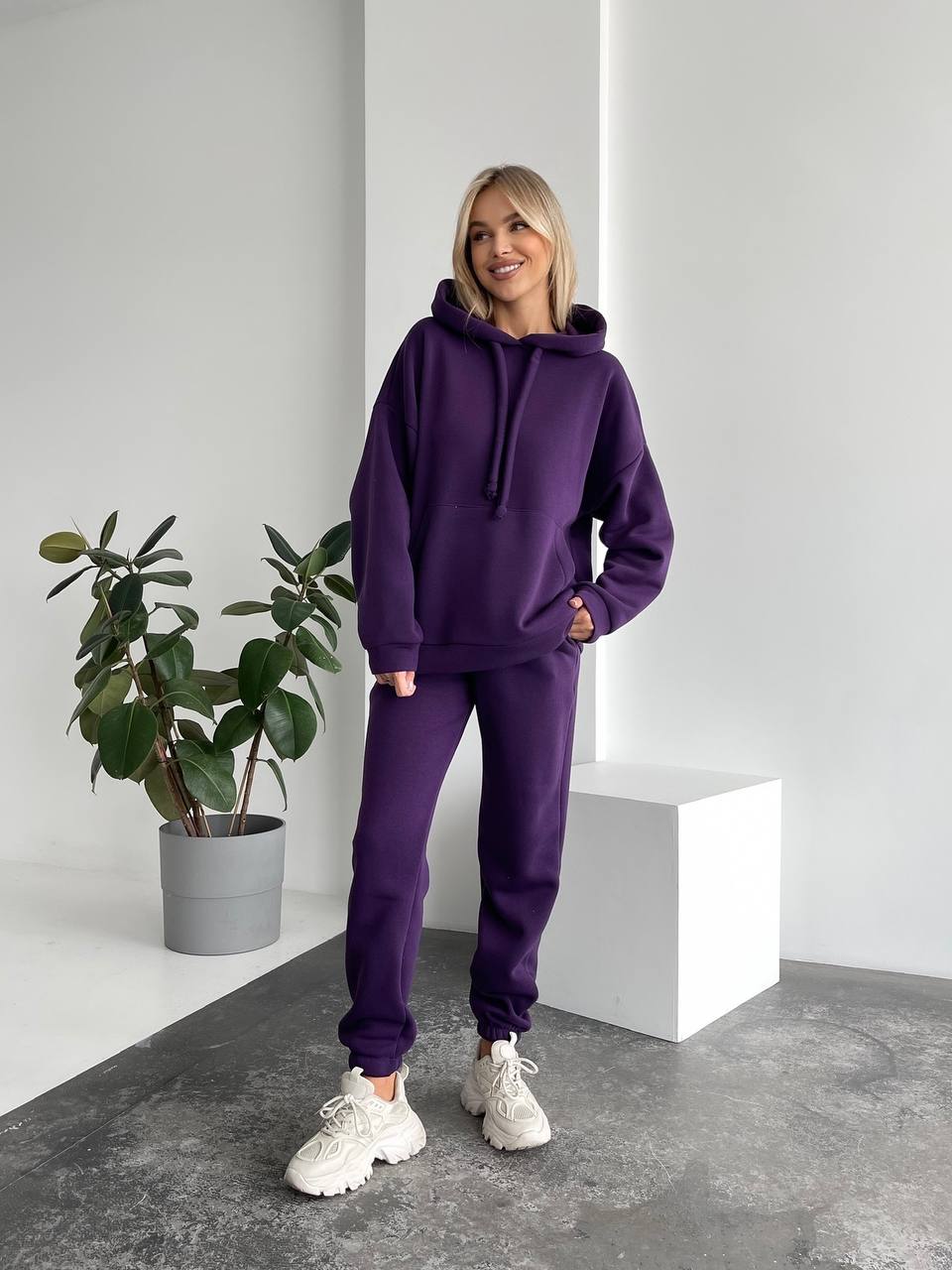 Person wearing a purple hoodie and pants standing in a minimalistic room with a plant.