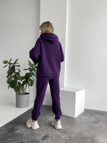 Person wearing a purple tracksuit standing in a minimalistic room with a plant.