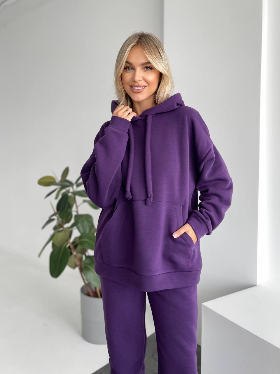 Woman wearing a purple hoodie and pants in a minimalistic room with a plant.
