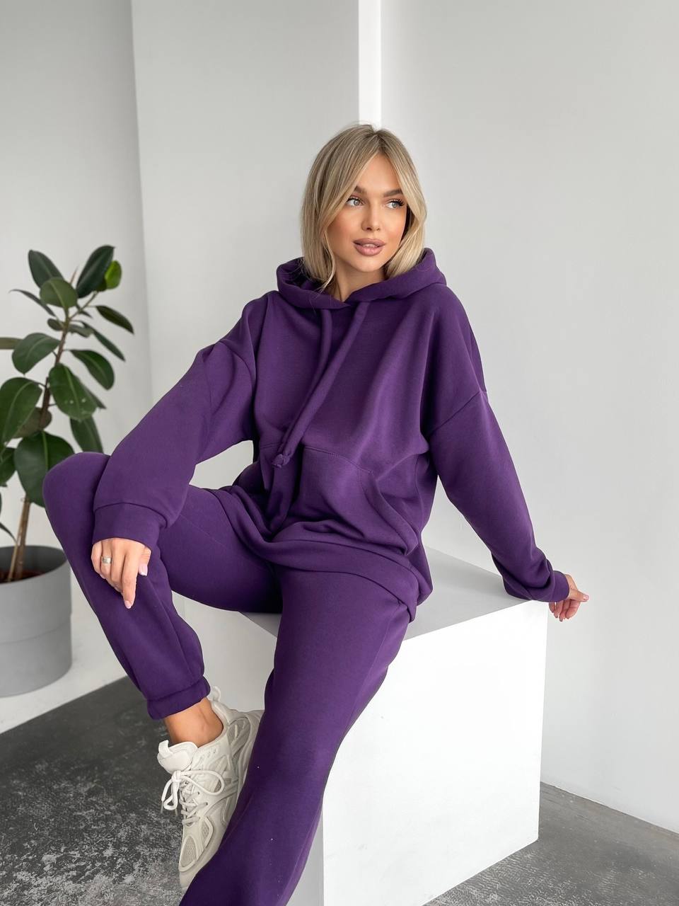 Woman wearing a purple tracksuit sitting on a white block with a plant in the background