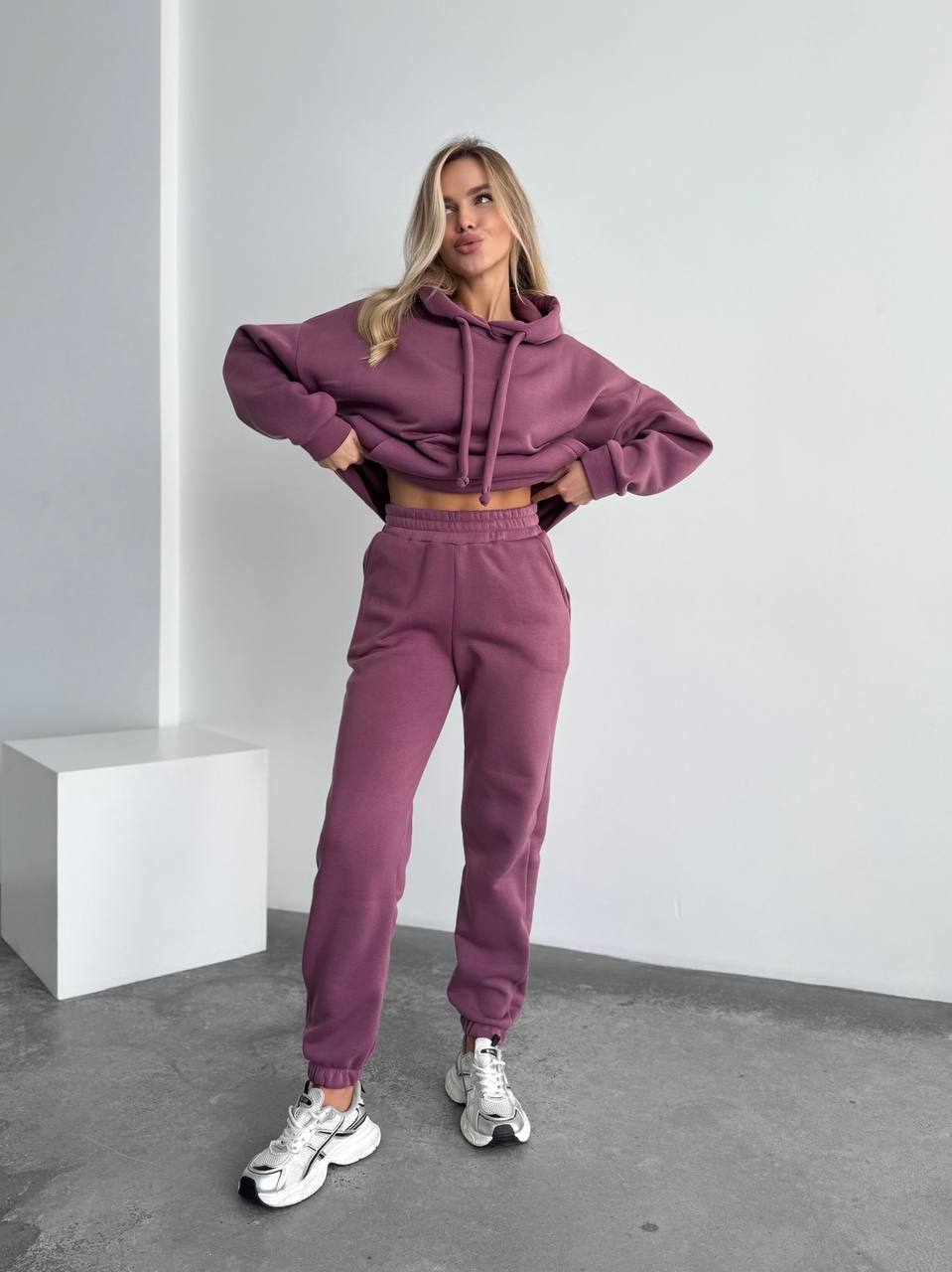 Woman wearing a purple tracksuit standing in a minimalistic room.