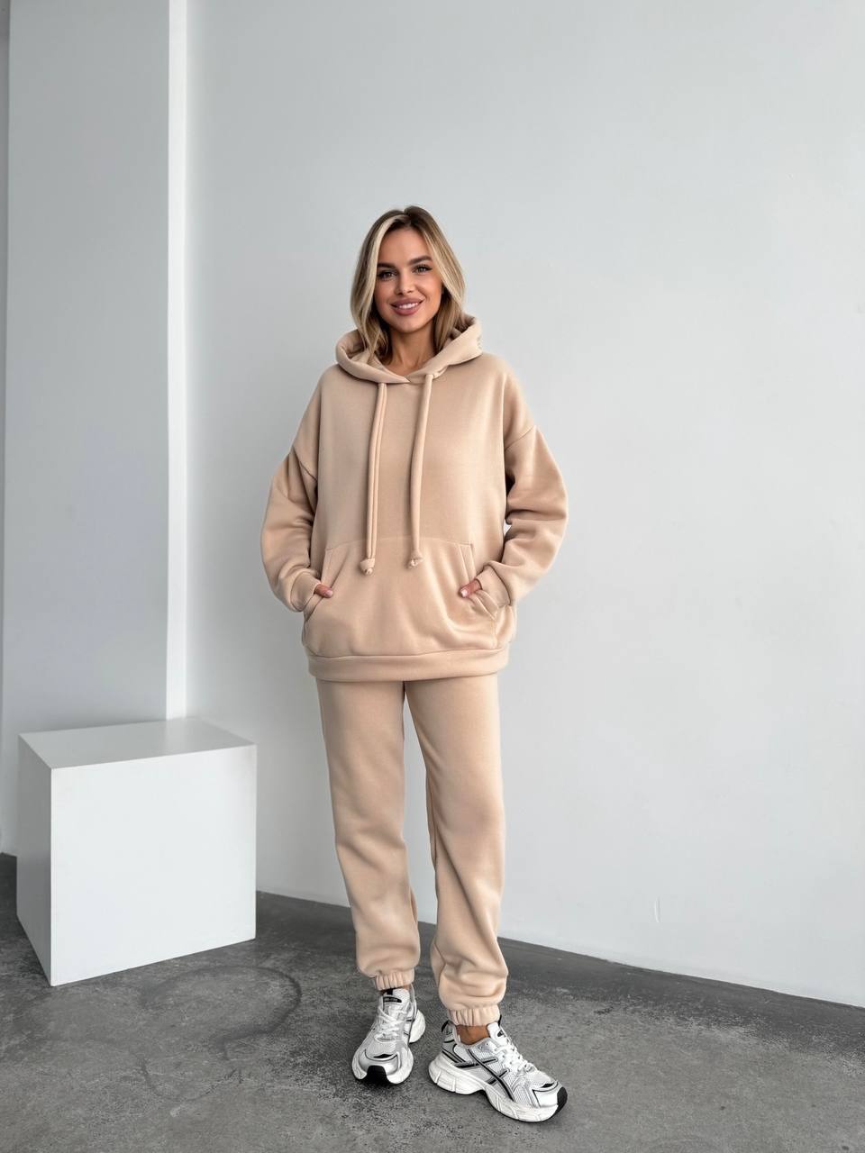 Woman wearing a beige tracksuit standing in a minimalistic room.