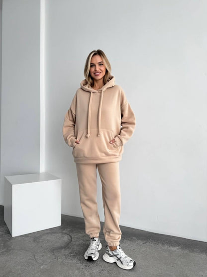 Woman wearing a beige tracksuit standing in a minimalistic room.