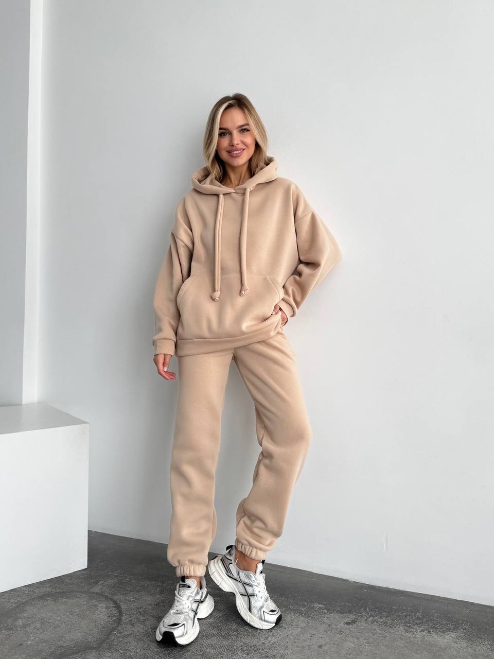 Woman wearing a beige tracksuit standing against a white wall.