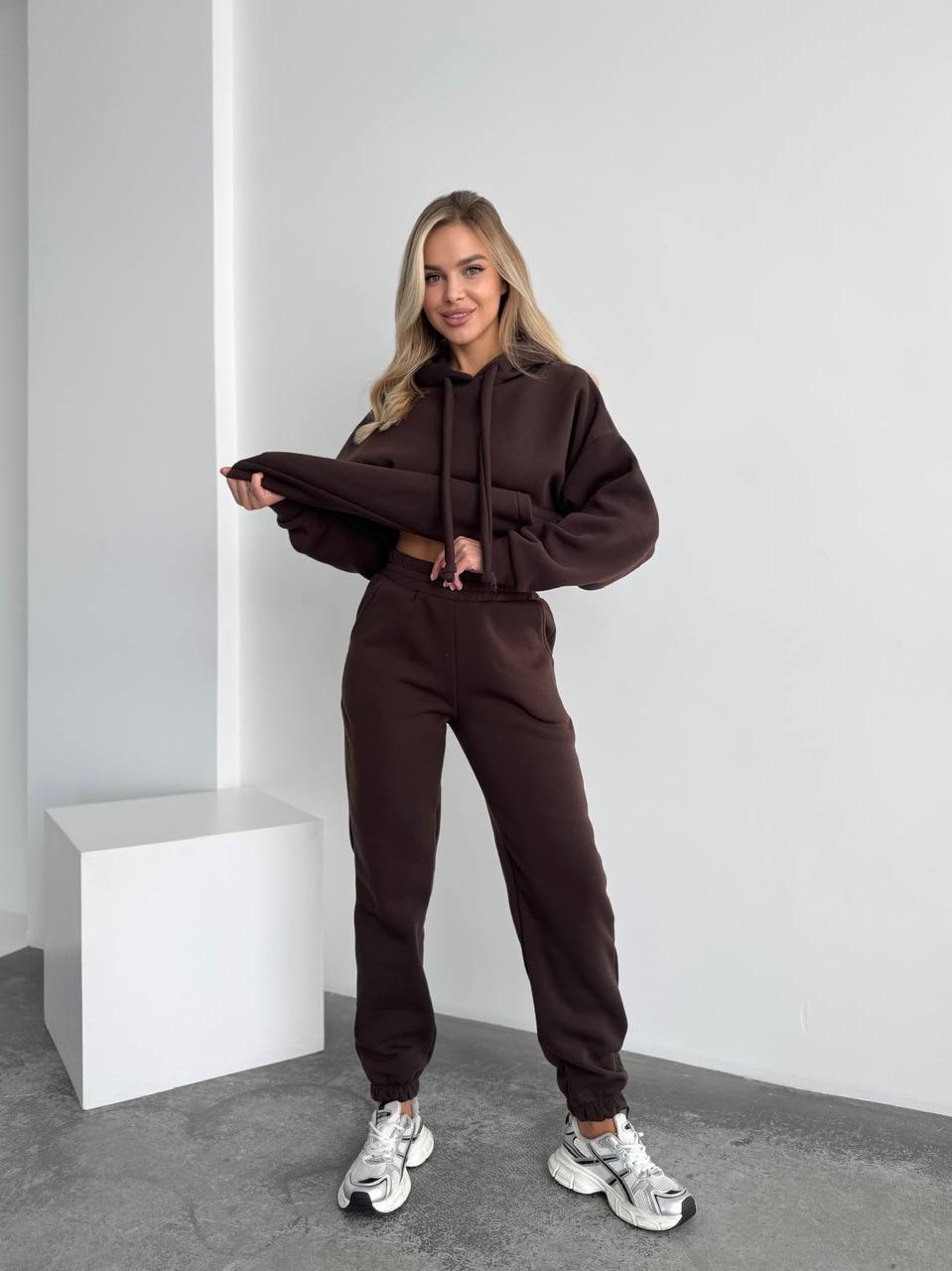 Woman wearing a brown tracksuit standing against a white wall.