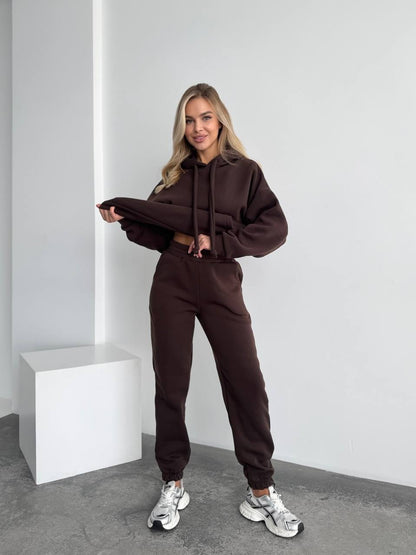 Woman wearing a brown tracksuit standing against a white wall.