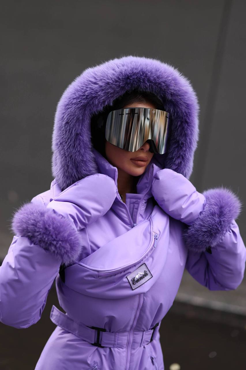 Person wearing a purple snowsuit with a fur-lined hood and goggles against a dark background