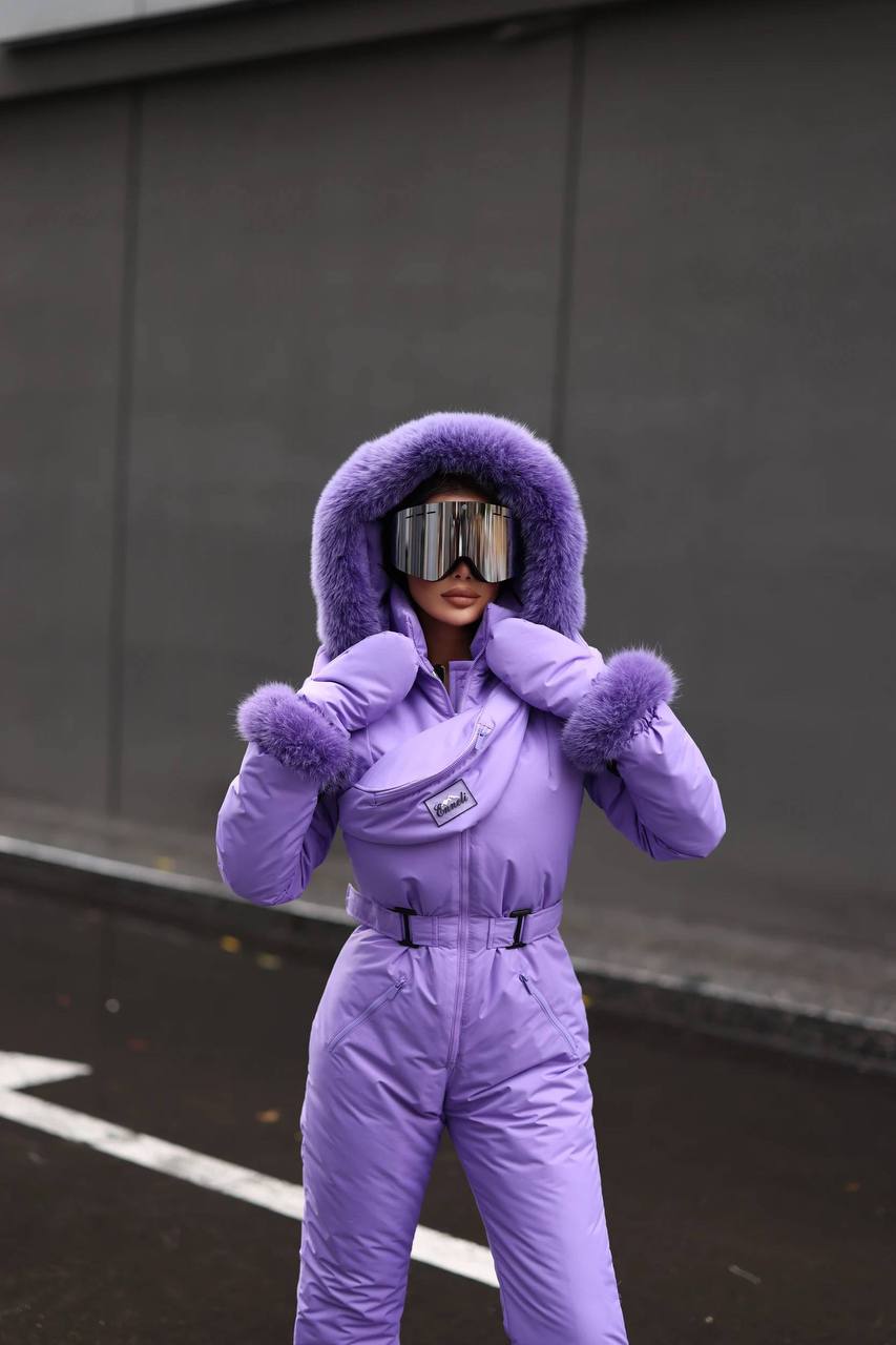 Person wearing a purple snowsuit with goggles in front of a dark wall.