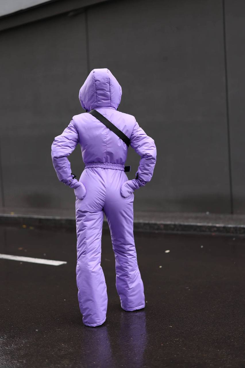 Person wearing a purple snowsuit standing on a wet pavement with a dark wall in the background.