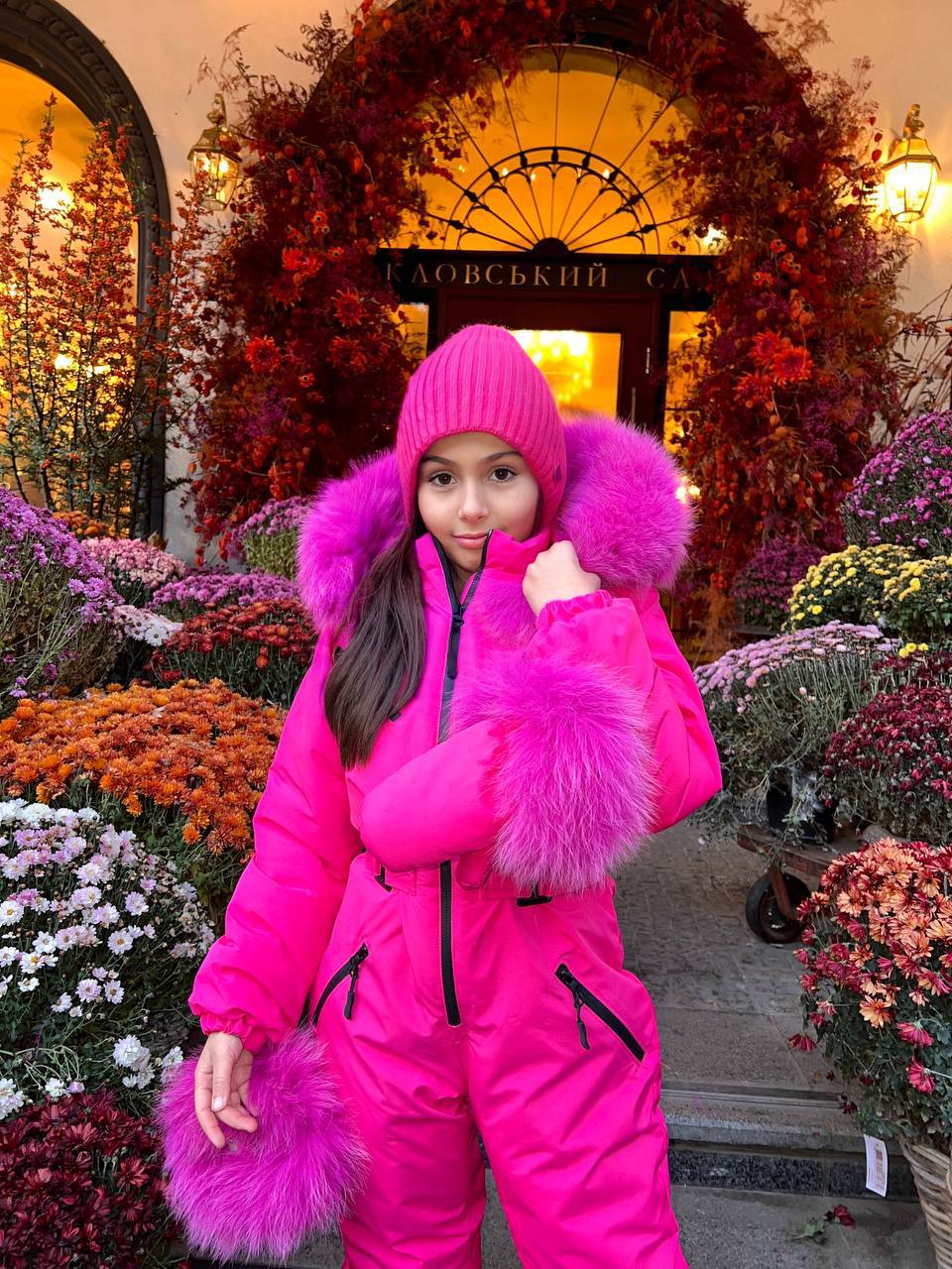Person wearing a bright pink snowsuit with fur trim in front of a decorative entrance.