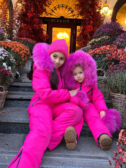 Two children in bright pink snowsuits with fluffy hoods sitting on steps surrounded by decorative flowers.