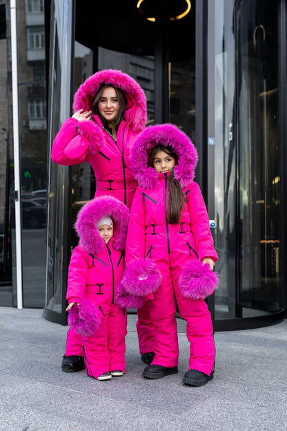 Family of four in matching pink snow outfits with fluffy hoods standing in front of a glass door.