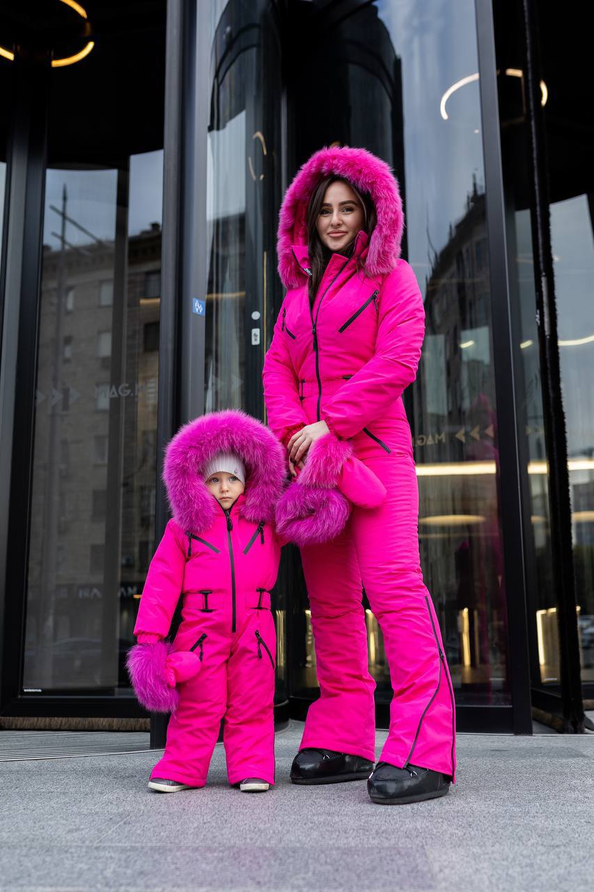 Two people in matching bright pink snowsuits standing in front of a modern building.