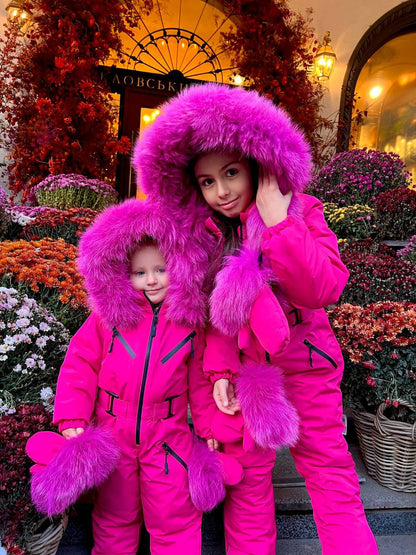 Two children in bright pink snow suits with fur hoods standing in front of a decorative entrance.
