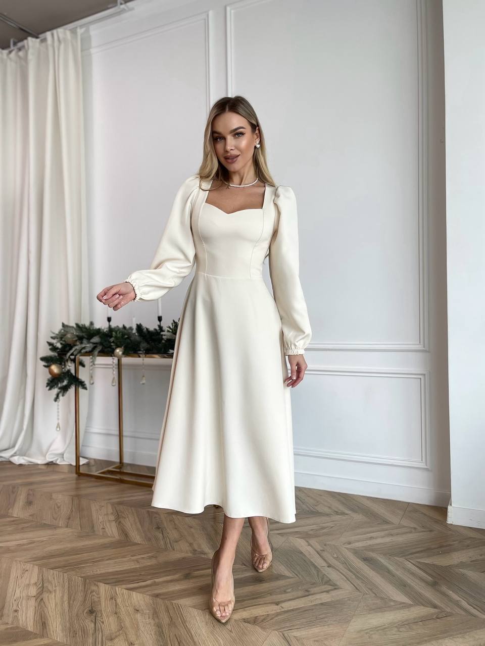 Woman in a white dress standing in a room with white walls and wooden floor.