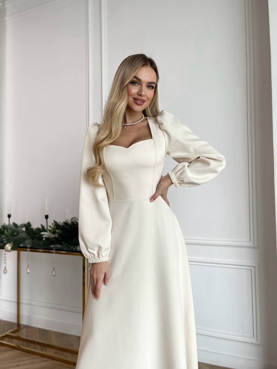 Woman in a white dress with a long, elegant coat standing in a stylish interior setting.