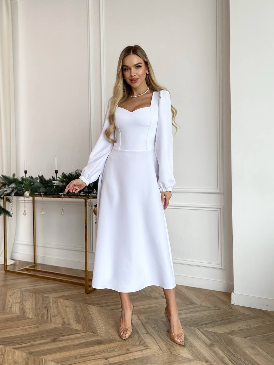 Woman in a white dress standing in a room with a decorative table and wall.