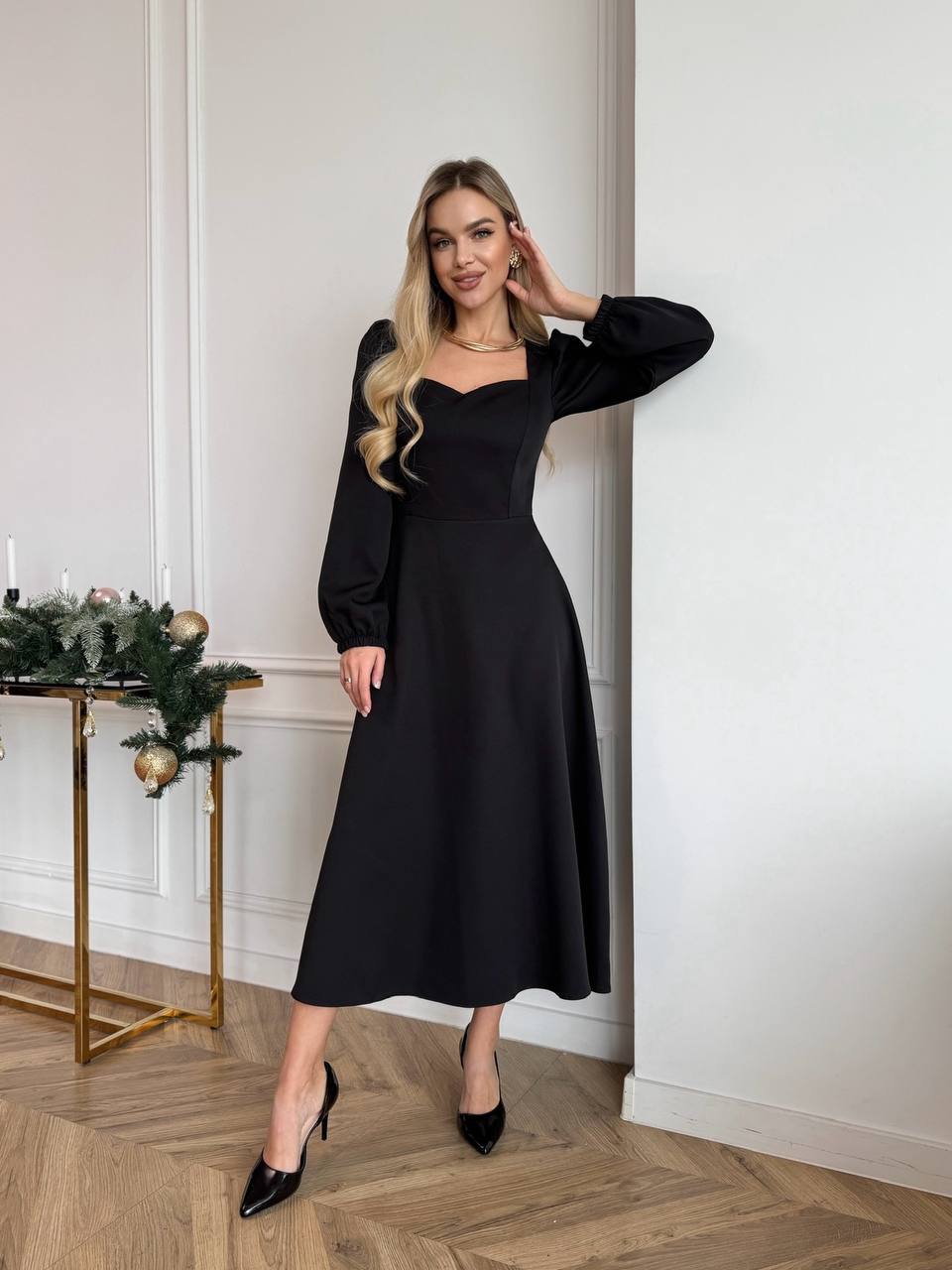 Woman in a black dress standing in a room with a decorative table and wall.