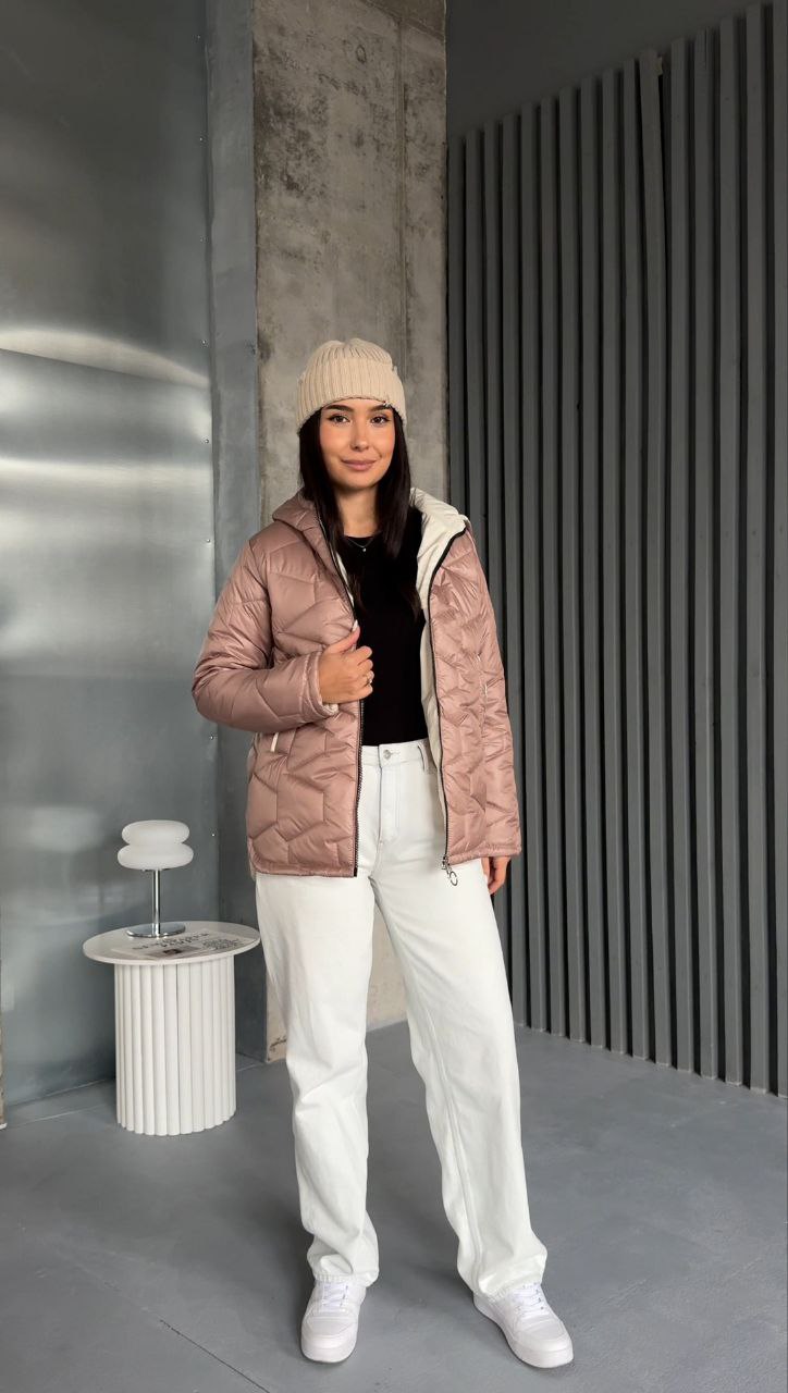 Person wearing a pink jacket and white pants standing indoors against a gray wall.