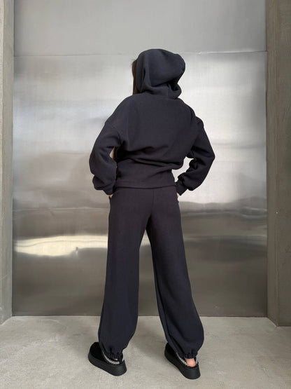 Person wearing a black hoodie and pants standing against a metallic wall.
