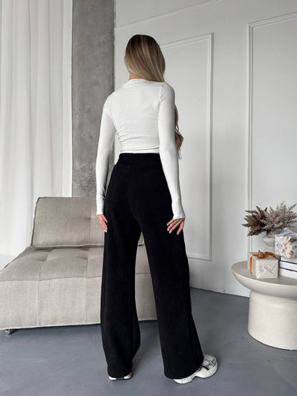 Person wearing a white long-sleeve top and black wide-leg pants in a minimalistic room.