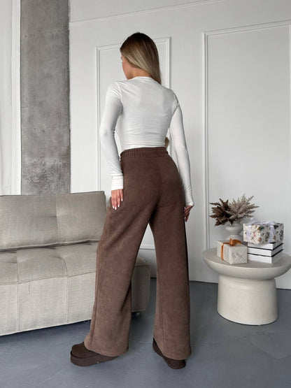 Person wearing a white long-sleeve top and brown wide-leg pants in a modern living room.