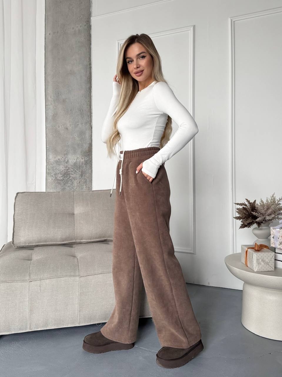 Woman wearing a white top and brown wide-leg pants in a modern living room.