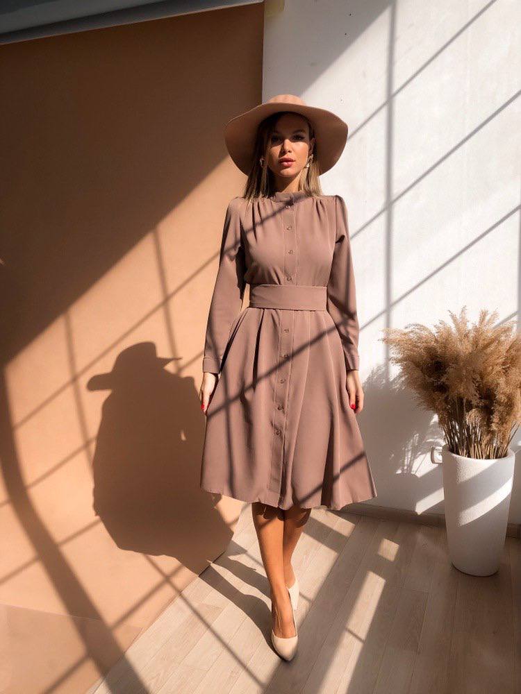 Woman in a brown dress and hat standing in sunlight casting shadows on a wall.