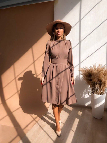 Woman in a brown dress and hat standing in sunlight casting shadows on a wall.