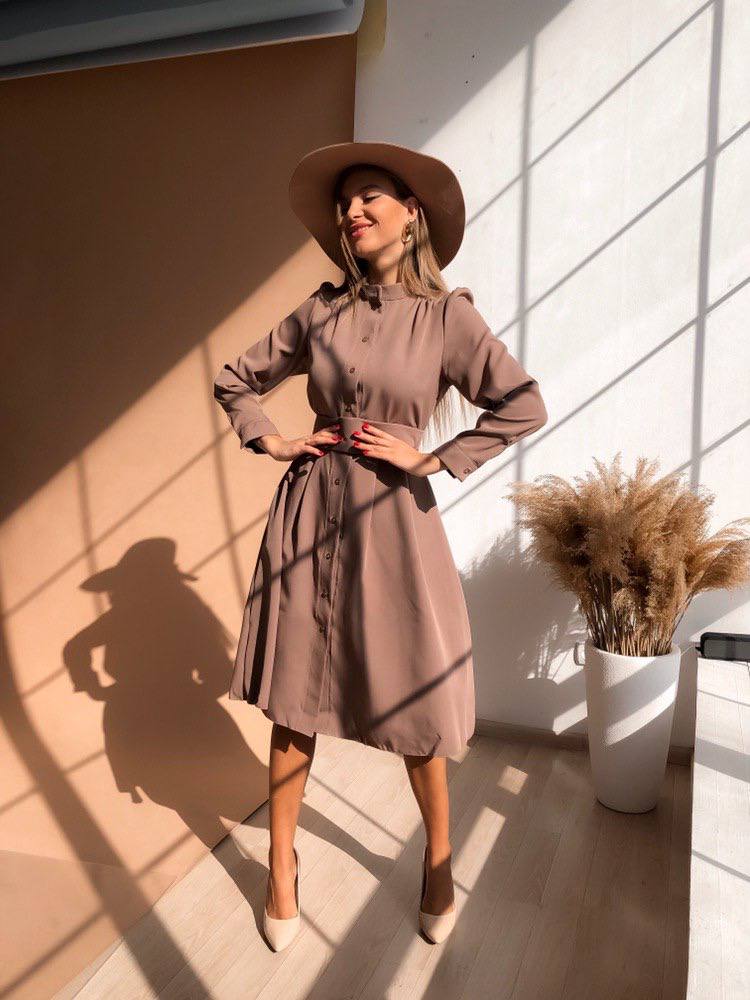 Woman in a beige dress and hat standing in a sunlit room with a plant in the background