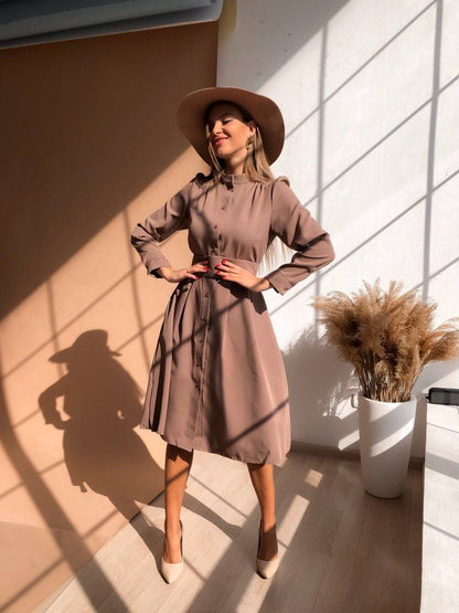 Woman in a beige dress and hat standing in a sunlit room with a plant in the background