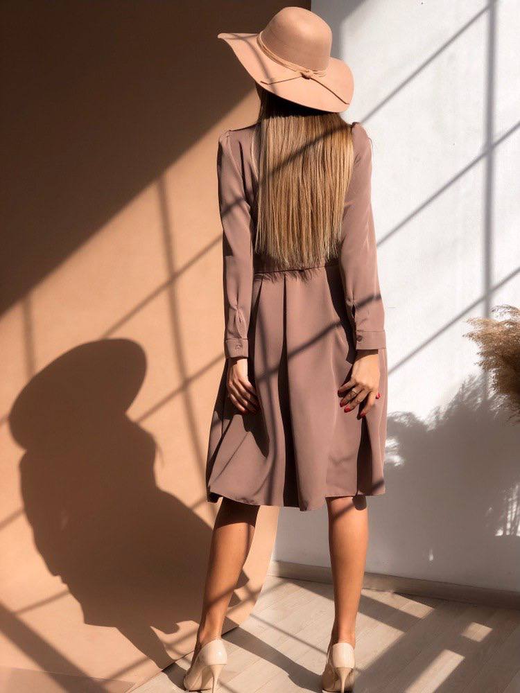 Woman wearing a long, light brown coat and wide-brimmed hat standing in a sunlit room.