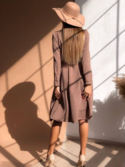 Woman wearing a long, light brown coat and wide-brimmed hat standing in a sunlit room.