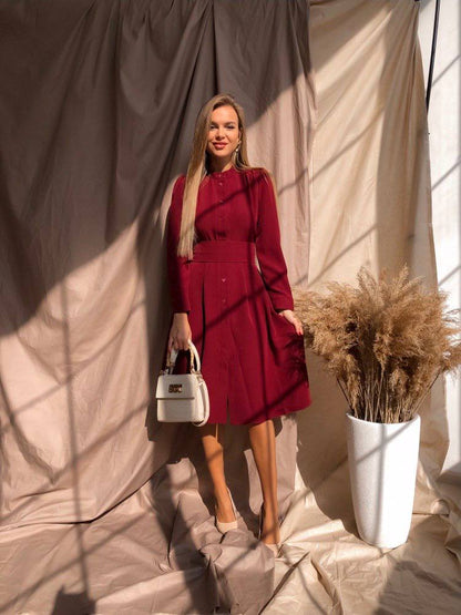 Woman in a red dress holding a white handbag, standing against a neutral backdrop with a plant.