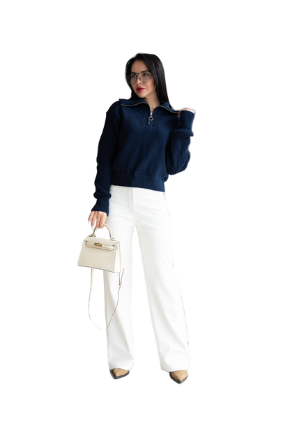 Woman wearing a navy blue sweater and white pants, holding a beige handbag on a white background