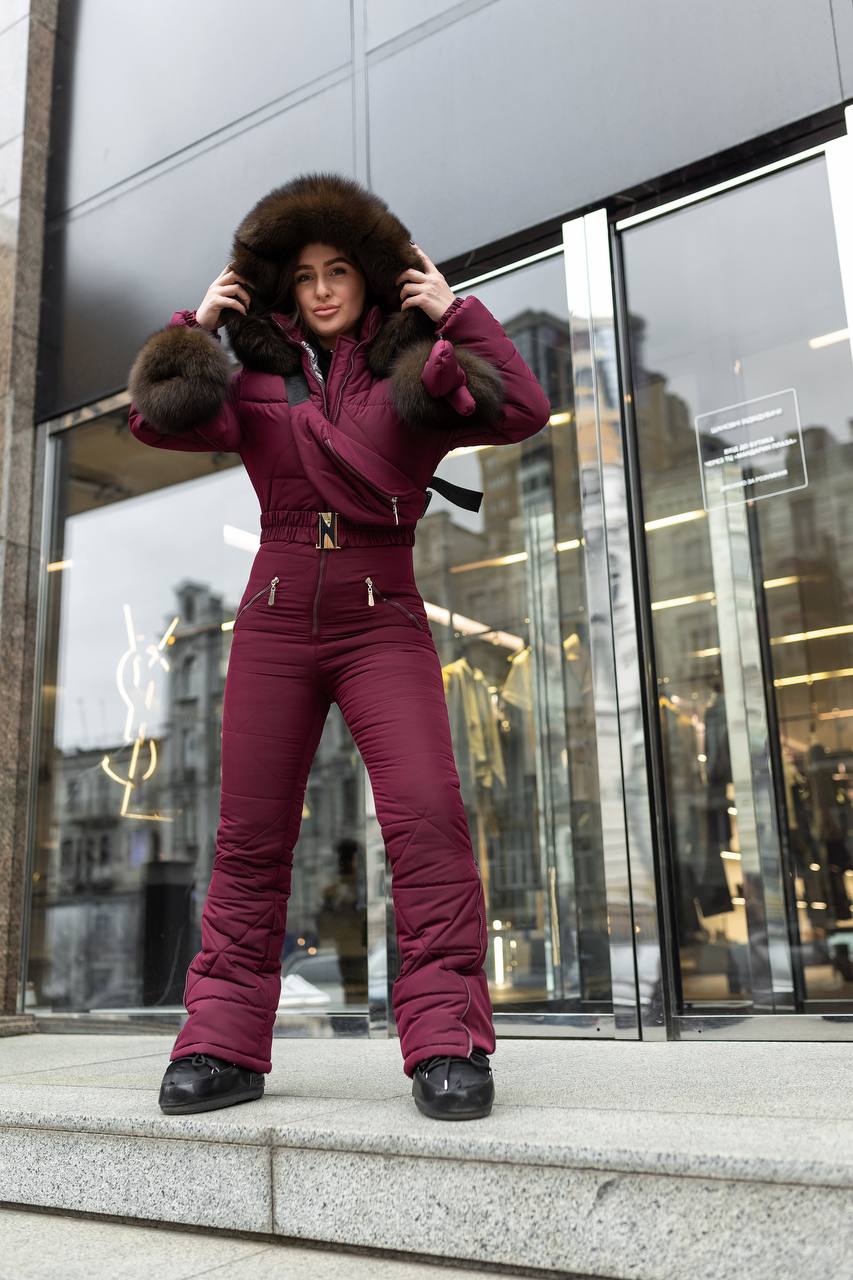 Person wearing a maroon snowsuit with fur trim standing in front of a modern building.