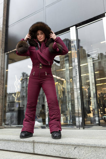 Person wearing a maroon snowsuit with fur trim standing in front of a modern building.