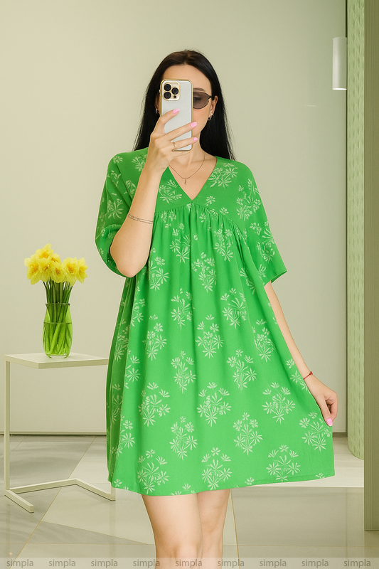 Woman wearing a green dress with white patterns, standing in a room with a vase of yellow flowers on a table.