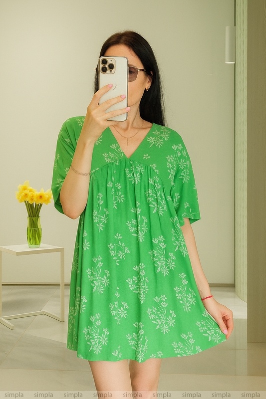 Woman taking a mirror selfie wearing a green floral dress in a room with a vase of flowers on a table.